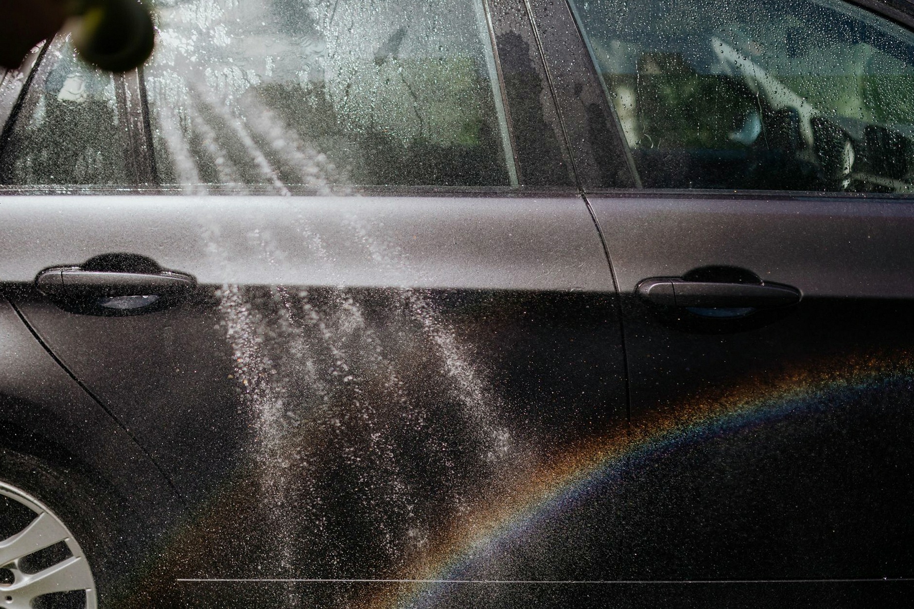 Close-up of a car being washed with a visible rainbow in sunlight.